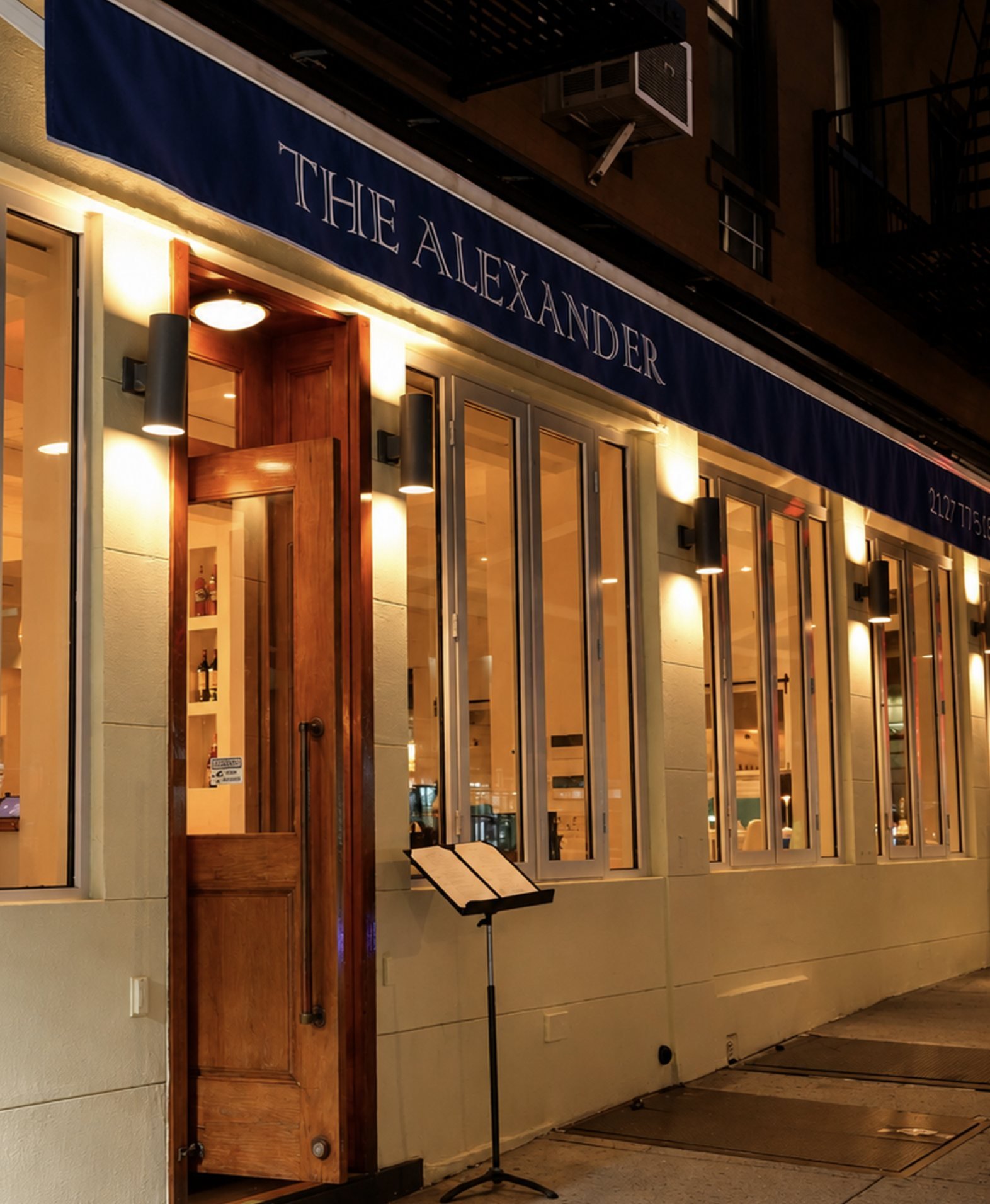 The Alexander at night: navy awning, warm-lit windows and an open wooden door